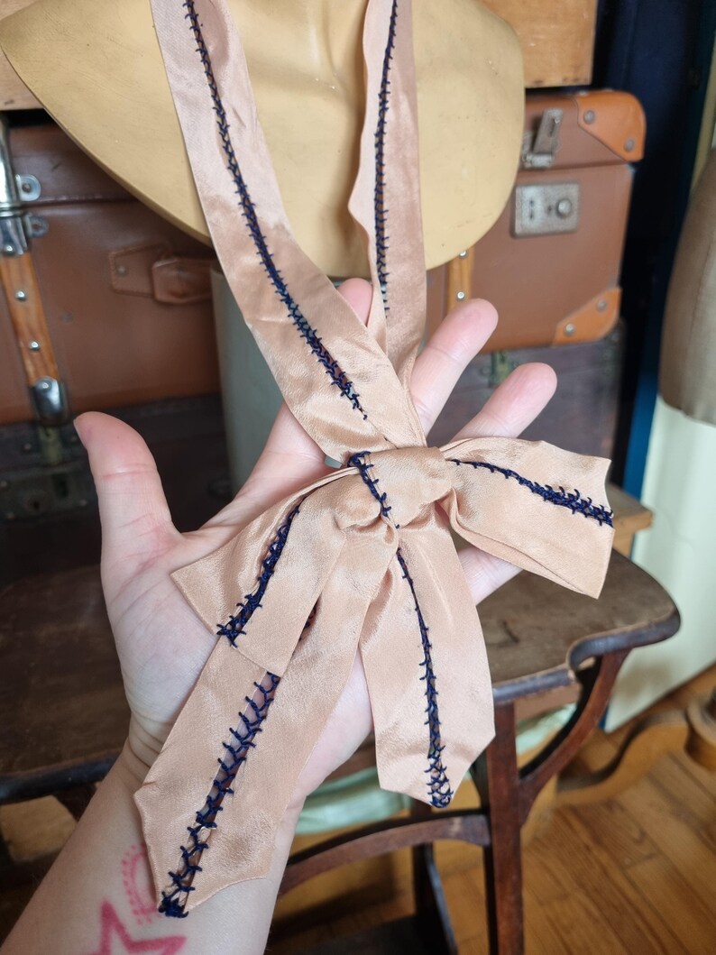 May include: A peach-colored satin ribbon with dark blue stitching detail. The ribbon is tied into a bow. The ribbon is likely vintage and is displayed against a vintage background.