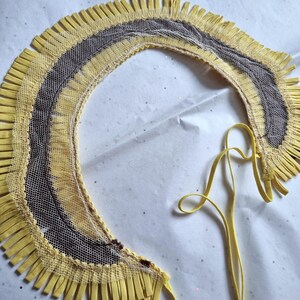 May include: A yellow and brown decorative collar with a fringe trim. The collar has a net-like center and a yellow ribbon tie. The design features a layered effect with a mix of textures and colors.