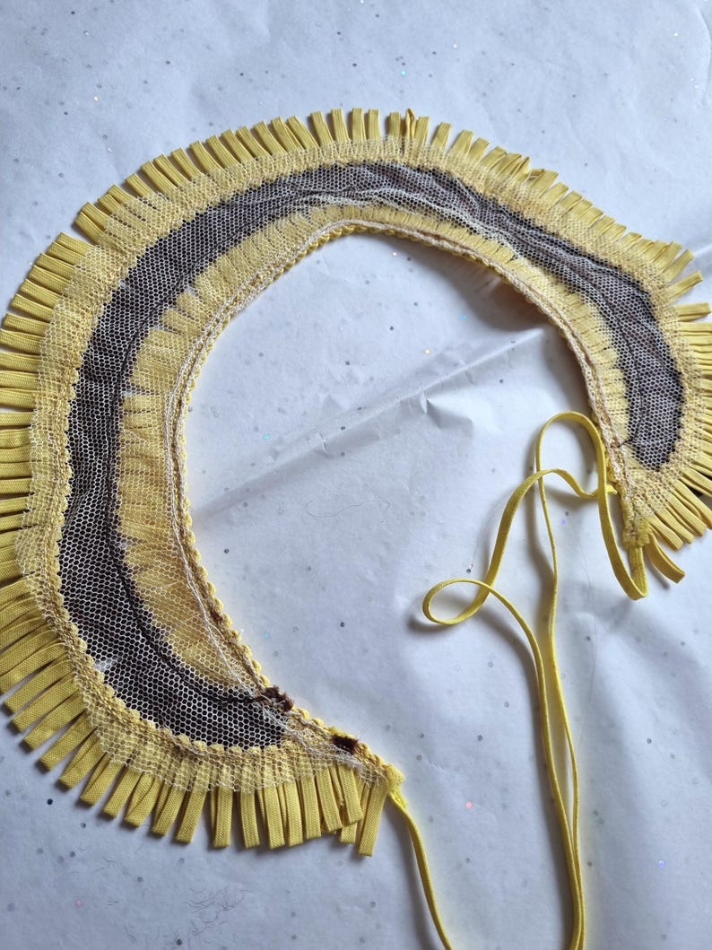 May include: A yellow and brown decorative collar with a fringe trim. The collar has a net-like center and a yellow ribbon tie. The design features a layered effect with a mix of textures and colors.