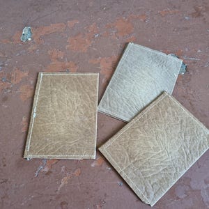 May include: Three rectangular, leather-effect coasters in shades of tan and beige are arranged on a distressed, reddish-brown surface. Each coaster has a stitched border in a lighter colour, adding a subtle contrast to the textured material.