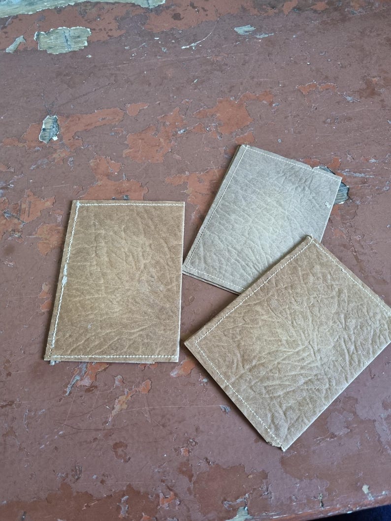 May include: Three rectangular, leather-effect coasters in shades of tan and beige are arranged on a distressed, reddish-brown surface. Each coaster has a stitched border in a lighter colour, adding a subtle contrast to the textured material.