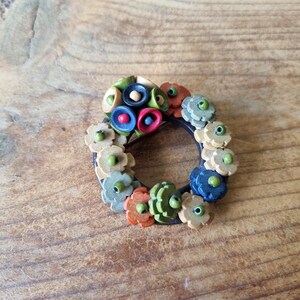 Peut inclure: Une broche florale artisanale avec une base circulaire marron foncé. La broche présente une variété de fleurs colorées en trois dimensions dans des tons de vert, orange, bleu et beige. La broche est posée sur un fond en bois.