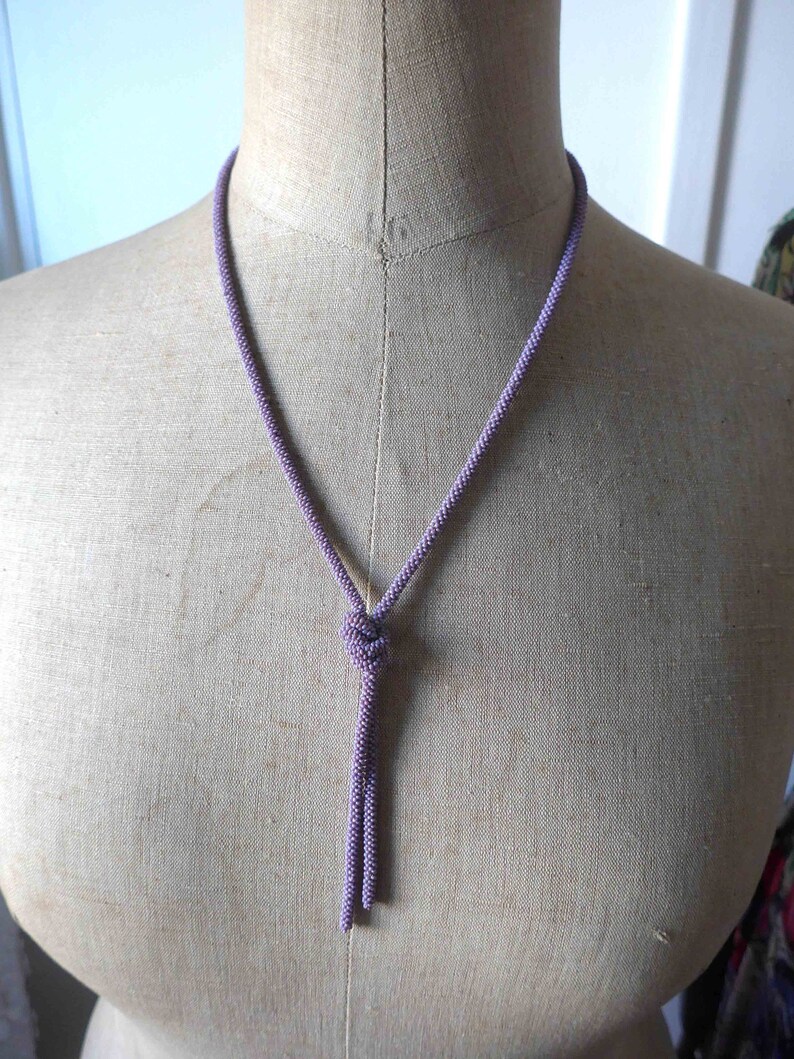 Pu&ograve; includere: Una collana di perline viola con un pendente annodato. La collana &egrave; esposta su un manichino rivestito di tessuto.