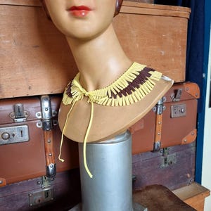 May include: A vintage mannequin head with a yellow and brown fringed collar. The collar is tied with a yellow string. The mannequin is displayed on a silver stand, with antique luggage in the background.