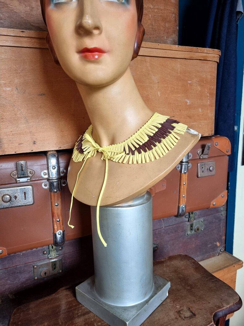 May include: A vintage mannequin head with a yellow and brown fringed collar. The collar is tied with a yellow string. The mannequin is displayed on a silver stand, with antique luggage in the background.