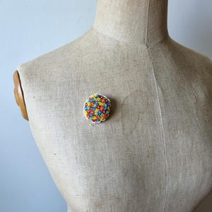 May include: A colorful beaded brooch with a circular design. The brooch is made of small, multi-colored beads in shades of blue, red, yellow, and green. The beads are arranged in a geometric pattern, creating a visually appealing design.