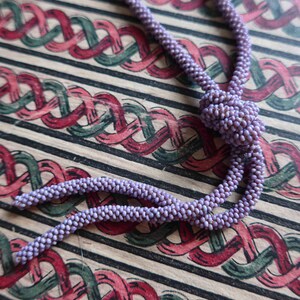 Pu&ograve; includere: Una collana di perline viola con un pendente annodato. La collana &egrave; appoggiata su un tessuto a motivi rossi e verdi.