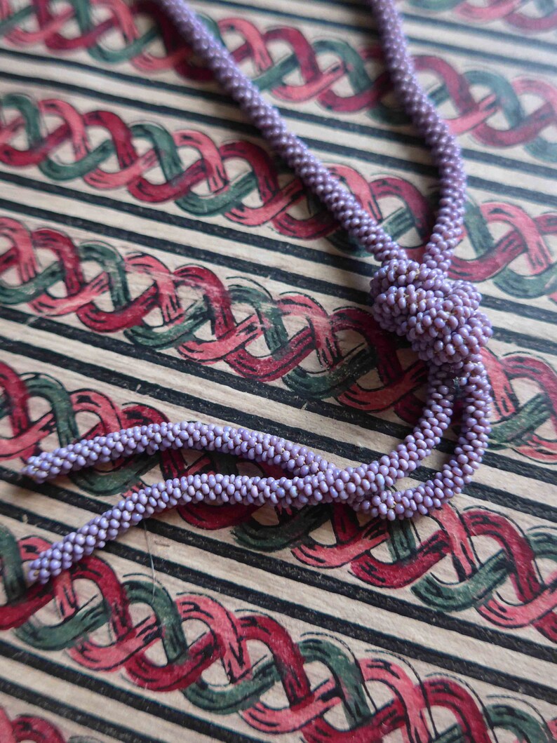 Pu&ograve; includere: Una collana di perline viola con un pendente annodato. La collana &egrave; appoggiata su un tessuto a motivi rossi e verdi.