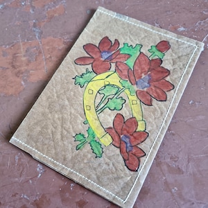 Puede incluir: Una tarjeta rectangular de color marr&oacute;n claro con un dise&ntilde;o dibujado a mano. El dise&ntilde;o presenta una herradura amarilla con un ramo de flores rojas y hojas verdes. La tarjeta est&aacute; cosida con hilo blanco alrededor de los bordes.