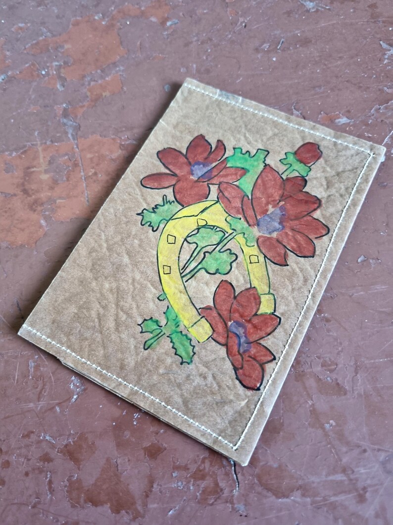 Puede incluir: Una tarjeta rectangular de color marr&oacute;n claro con un dise&ntilde;o dibujado a mano. El dise&ntilde;o presenta una herradura amarilla con un ramo de flores rojas y hojas verdes. La tarjeta est&aacute; cosida con hilo blanco alrededor de los bordes.