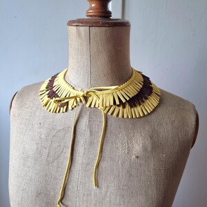 May include: A decorative collar with a fringed design. The collar features alternating sections of yellow and brown fringe. It is tied with yellow cords and displayed on a mannequin.