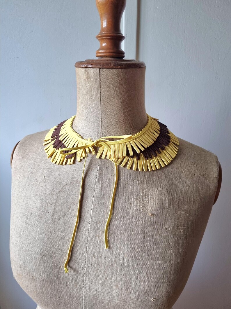 May include: A decorative collar with a fringed design. The collar features alternating sections of yellow and brown fringe. It is tied with yellow cords and displayed on a mannequin.