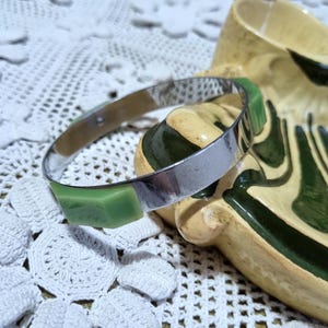 May include: A silver bangle bracelet with green plastic accents. The bracelet is resting on a ceramic dish with green and white designs.