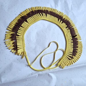 May include: A handmade, yellow and brown fringed collar with a tie closure. The collar is made of layered, cut strips of paper or fabric, creating a textured, decorative effect. The design features alternating sections of yellow and brown fringe.