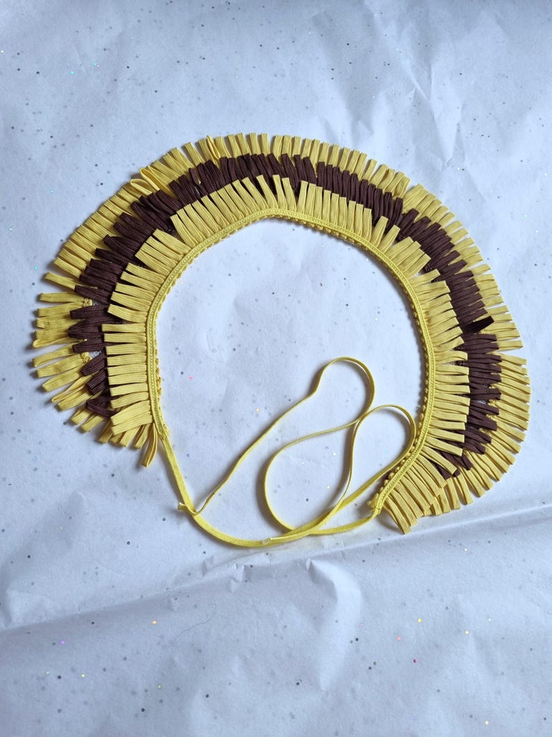 May include: A handmade, yellow and brown fringed collar with a tie closure. The collar is made of layered, cut strips of paper or fabric, creating a textured, decorative effect. The design features alternating sections of yellow and brown fringe.
