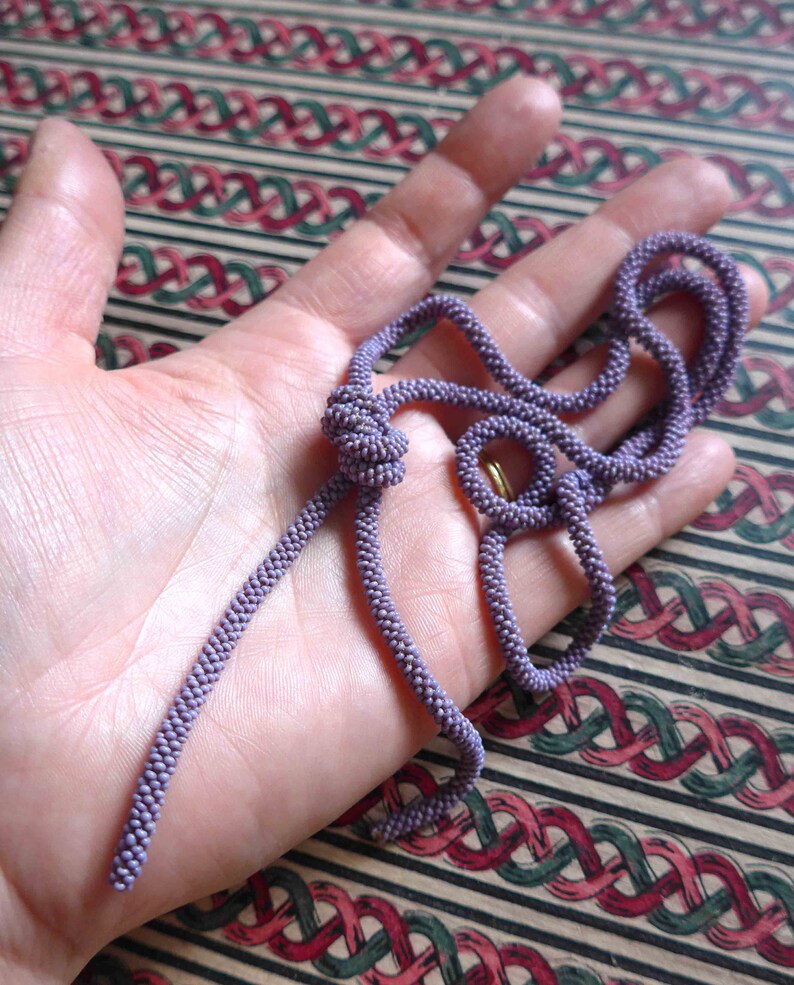 Pu&ograve; includere: Una lunga e sottile collana di perline viola con un designo annodato. La collana &egrave; fatta di piccole perline rotonde e si trova su una mano.