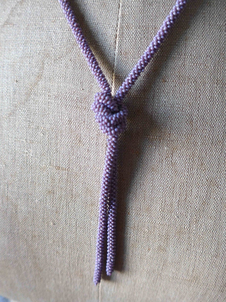 Pu&ograve; includere: Una collana lunga e sottile fatta di piccole perline viola. La collana &egrave; annodata in cima e pende in due fili.