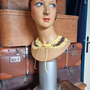 May include: A vintage mannequin head with painted features, including blue eyeshadow and red lipstick, wears a yellow and brown fringed collar. The mannequin is mounted on a silver stand, with antique luggage in the background.