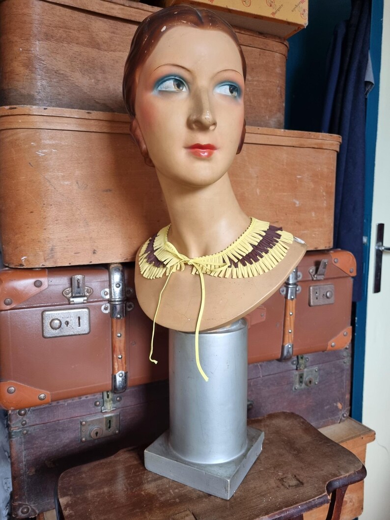 May include: A vintage mannequin head with painted features, including blue eyeshadow and red lipstick, wears a yellow and brown fringed collar. The mannequin is mounted on a silver stand, with antique luggage in the background.