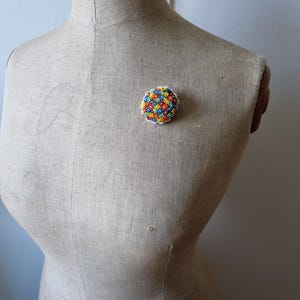 May include: A colorful beaded brooch with a round shape and a white background. The beads are arranged in a flower pattern and are in shades of red, orange, yellow, green, blue, and purple.