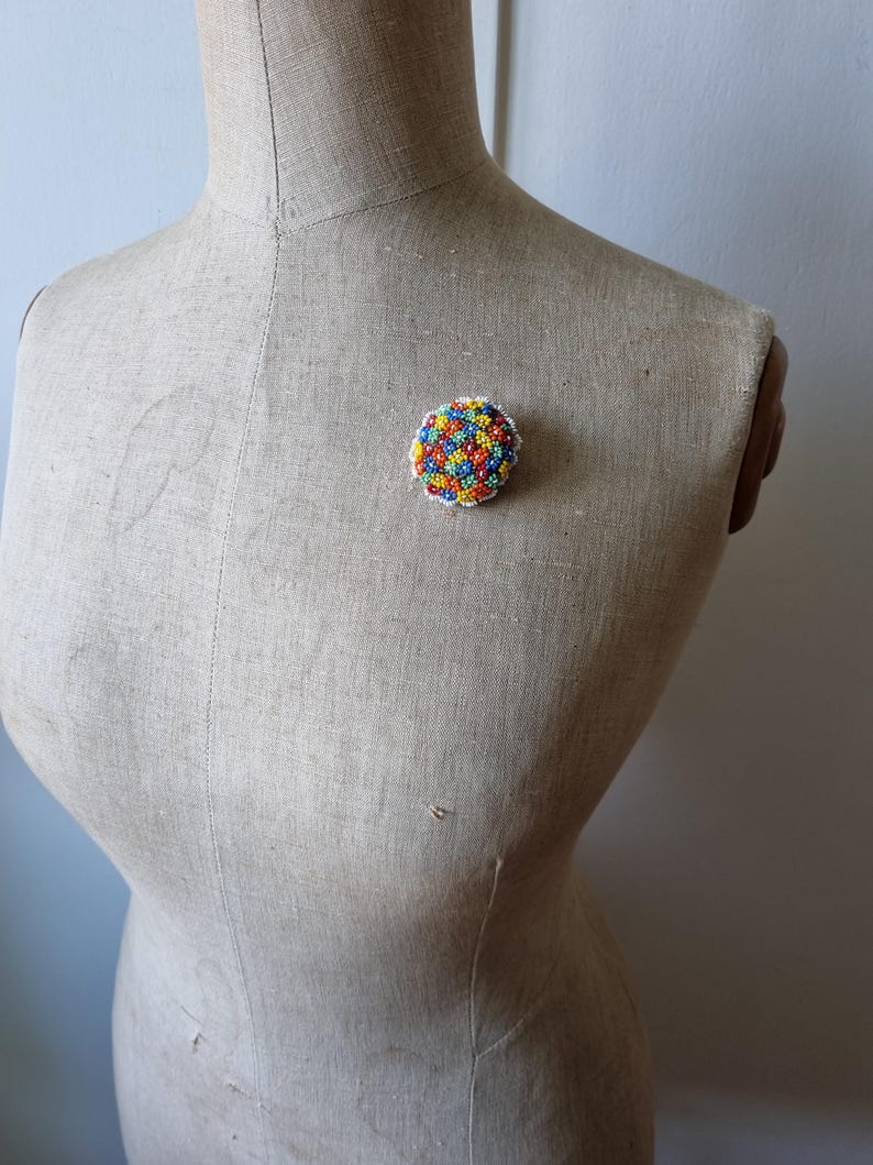 May include: A colorful beaded brooch with a round shape and a white background. The beads are arranged in a flower pattern and are in shades of red, orange, yellow, green, blue, and purple.