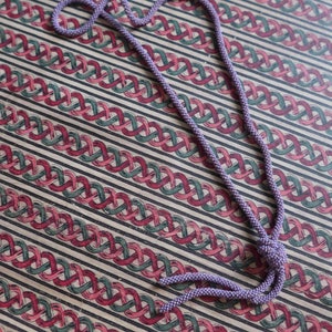 Pu&ograve; includere: Una lunga collana di perline viola con un'estremit&agrave; annodata. La collana &egrave; appoggiata su una superficie a motivi.