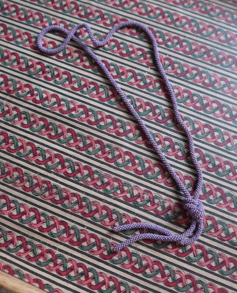 Pu&ograve; includere: Una lunga collana di perline viola con un'estremit&agrave; annodata. La collana &egrave; appoggiata su una superficie a motivi.