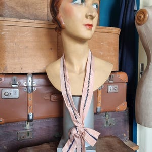 May include: A vintage mannequin head with painted features, wearing a light pink ribbon with dark blue stitching. The mannequin is displayed on a metal stand, with antique luggage and wooden boxes in the background.