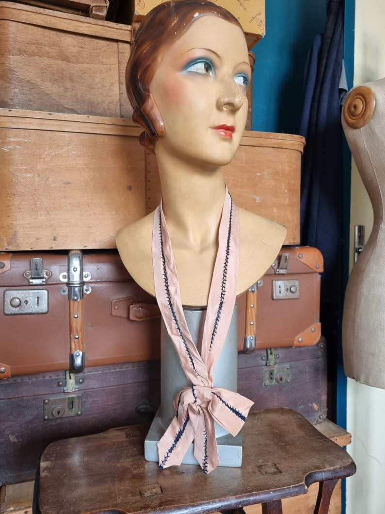 May include: A vintage mannequin head with painted features, wearing a light pink ribbon with dark blue stitching. The mannequin is displayed on a metal stand, with antique luggage and wooden boxes in the background.