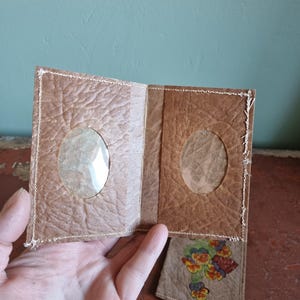 May include: Handmade brown paper photo album with two clear oval windows. The album is stitched with white thread and has a butterfly design on the bottom right corner. The album is open, revealing the inside pages.