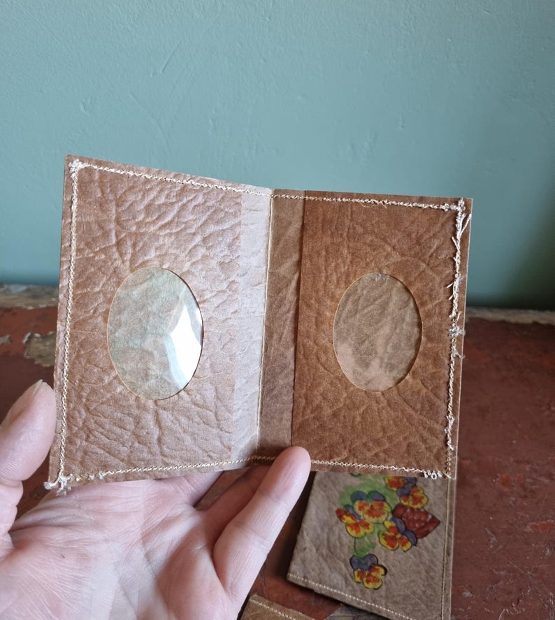 May include: Handmade brown paper photo album with two clear oval windows. The album is stitched with white thread and has a butterfly design on the bottom right corner. The album is open, revealing the inside pages.
