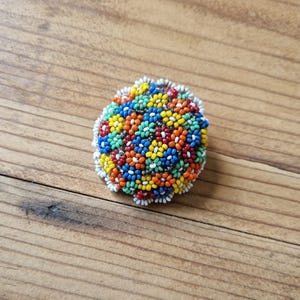 May include: A colorful beaded brooch with a floral design. The brooch is made of small, round beads in shades of red, orange, yellow, green, blue, and white.