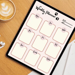 Op de afbeelding: Een digitale weekplanner op een tablet met een lichtroze achtergrond en een rasterindeling. De planner bevat secties voor elke dag van de week, doelen en notities. De tekst "Weekly Planner" staat bovenaan, met pootafdrukken. Op de achtergrond staat een kopje koffie.