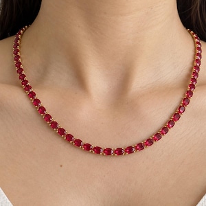 Può includere: Una collana tennis color oro caratterizzata da una fila continua di vivaci pietre preziose rosse dal taglio ovale, fissate con griffe. La collana poggia attorno al collo, mostrando un design uniforme con maglie in metallo lucido.