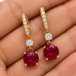 Peut inclure: Une paire de boucles d'oreilles pendantes en or avec une pierre précieuse ronde rouge foncé suspendue à une pierre ronde transparente plus petite. Les crochets sont ornés d'une rangée de minuscules pierres transparentes et scintillantes.