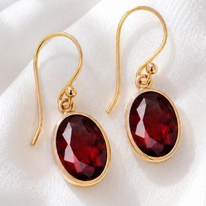 Puede incluir: Un par de pendientes colgantes de oro con piedras preciosas de granate rojo oscuro facetadas en forma ovalada. Cada pendiente cuelga de un gancho de alambre de oro curvado, mostrando un engaste de oro pulido que enmarca la piedra roja.