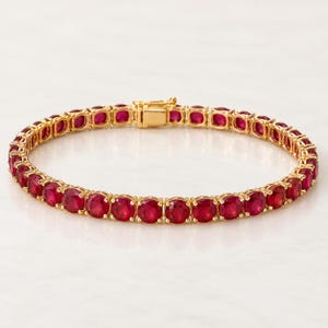 May include: A gold tennis bracelet featuring a continuous row of round, vibrant red rubies set in polished gold metal prongs. The bracelet includes a secure, rectangular gold box clasp mechanism that connects the two ends of the circular jewellery piece.