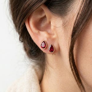 Peut inclure: Une paire de boucles d'oreilles à clous en or avec des pierres précieuses rouge foncé en forme de goutte. Chaque boucle est sertie dans un chaton en or lisse, mettant en valeur une teinte cramoisie vive dans une taille poire classique.