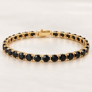 Könnte beinhalten: Ein goldfarbenes Tennisarmband mit einer durchgehenden Reihe runder, facettierter schwarzer Steine, die in einzelnen Metallfassungen sitzen. Das Armband verfügt über einen sicheren, rechteckigen Kastenschloss-Verschluss an der Oberseite.