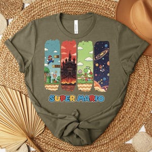 May include: Olive green t-shirt featuring a colorful Super Mario graphic. The design includes four vertical panels depicting scenes from the game, with the words "SUPER MARIO" in a blue and orange font.