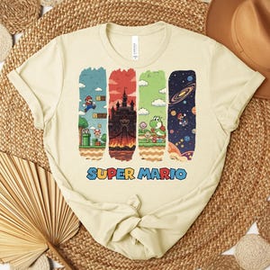 May include: Cream-colored t-shirt featuring a colorful Super Mario graphic. The design includes four vertical panels with scenes from the game, including Mario, a castle, Yoshi, and a space scene. The words "SUPER MARIO" are printed in blue.