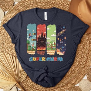 May include: Navy blue t-shirt featuring a colorful Super Mario graphic. The design includes four vertical panels with scenes from the game, including Mario, a castle, Yoshi, and space. The words "SUPER MARIO" are printed in a colorful font.