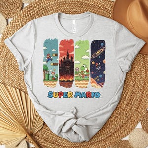 May include: A light gray t-shirt featuring a colorful Super Mario graphic. The design includes four vertical rectangular panels with scenes from the game, and the words "SUPER MARIO" in a colorful font.