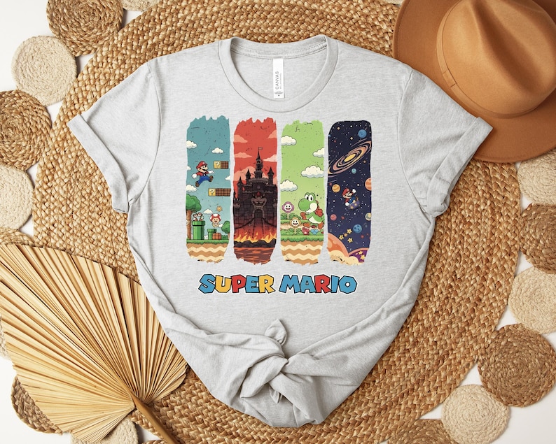 May include: A light gray t-shirt featuring a colorful Super Mario graphic. The design includes four vertical rectangular panels with scenes from the game, and the words "SUPER MARIO" in a colorful font.