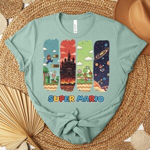 May include: A light green t-shirt featuring a Super Mario graphic. The graphic includes four vertical panels with scenes from the game, including Mario, a castle, and Yoshi. The words "SUPER MARIO" are printed in blue, red, and yellow.