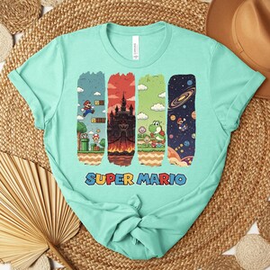 May include: A light teal t-shirt featuring a Super Mario graphic. The graphic includes four vertical panels with scenes from the game, including Mario, a castle, Yoshi, and space. The words "SUPER MARIO" are printed in colorful letters below the panels.