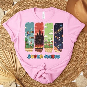 May include: Pink t-shirt featuring a colorful Super Mario graphic. The design includes four vertical panels depicting scenes from the game, with the words "Super Mario" in a blue and red font at the bottom. The shirt is made of a soft material.