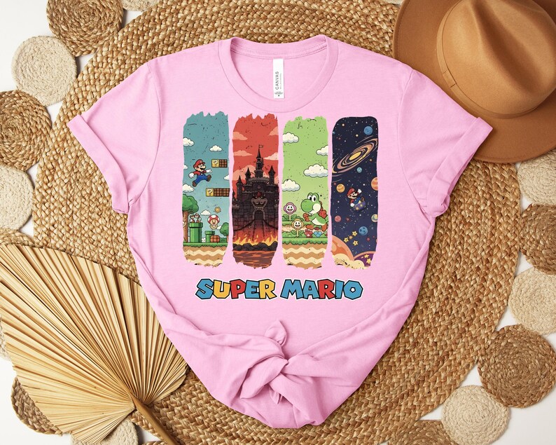 May include: Pink t-shirt featuring a colorful Super Mario graphic. The design includes four vertical panels depicting scenes from the game, with the words "Super Mario" in a blue and red font at the bottom. The shirt is made of a soft material.