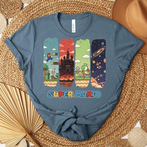 May include: A blue t-shirt featuring a Super Mario design. The graphic includes four vertical panels with scenes from the game, including Mario, a castle, Yoshi, and a space scene. The words "SUPER MARIO" are printed in colorful letters at the bottom.