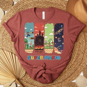 May include: A maroon t-shirt featuring a Super Mario graphic. The design includes four vertical panels with scenes from the game, including Mario, a castle, Yoshi, and a space scene. The words "SUPER MARIO" are printed in a colorful font.